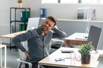 Mature businessman massaging aching neck and back, suffering from pain after computer work, sitting at workplace