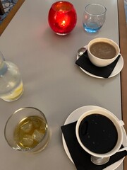 Obraz na płótnie Canvas Interior view of coffee drinks in white cups sat on black serviette napkin with saucers and spoons, vegan one black other oat milk, glass of brandy spirit with ice, jug lemon water flat lay on table