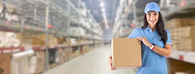 Young delivery woman in work clothes in front of a huge warehouse holding cardboard box for delivery to the customer