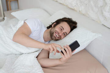 Smiling young european male with stubble wake up in bed, chatting on smartphone, reading news in bedroom