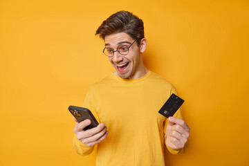 Cheerful dark haired man holds smartphone and credit card involved in online shopping purchases...