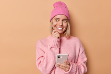 Pretty delighted woman looks awa with dreamy expression uses cellphone chats with friend posts something in social media wears pink jumper and hat poses over brown background thinks about mobile offer - obrazy, fototapety, plakaty