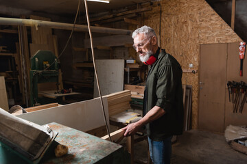 bearded carpenter holding wooden plank near thickness planer in workshop.