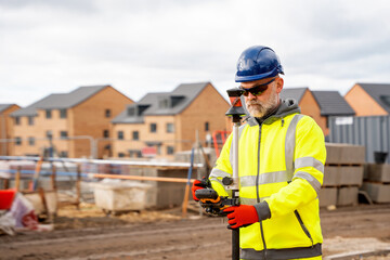 Site engineer in hi-viz working on house building construction site using modern surveying equipment against new houses nad blue cloudy sky background