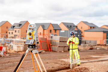 Site engineer in hi-viz working on house building construction site using modern surveying equipment against new houses nad blue cloudy sky background