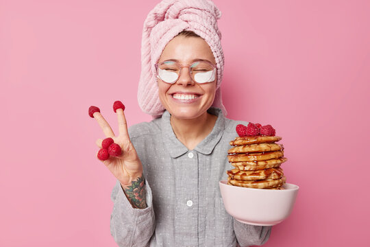 Positive young woman has fun going to eat yummy pancakes with raspberries and honey makes peace gesture smiles gladfully wears pajama and towel on head poses against pink background stays happy