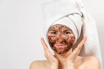 A young woman with a coffee mask on her face and a bath towel on her head smiles and looks into the camera on a white background, a place for text.