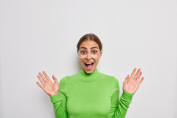Happy excited brunette European woman exclaims loudly keeps palms raised gets unexpected news wears...