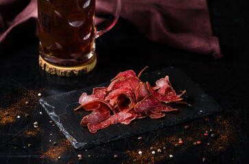An appetizing snack for beer is basturma, dried meat (pork or beef). Close-up of a delicious dried snack. Black slate board and beer in the background.