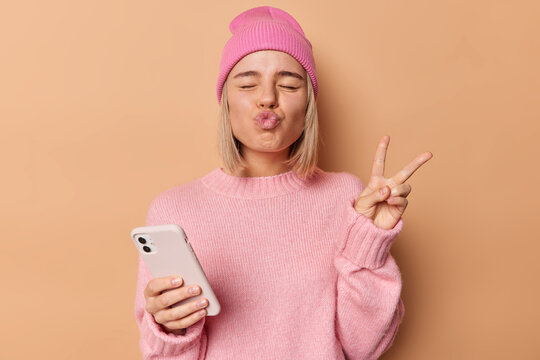 Funny woman keeps eyes closed lips folded makes peace gesture holds mobile phone enjoys online communication shows victory sign dressed in casual jumper and hat isolated over beige background.