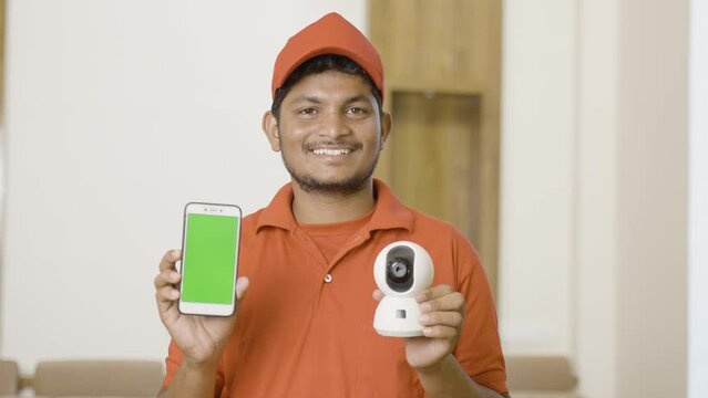 happy technician showing green screen mobile phone by holding smart cctv camera while looking camera at home - concept of technology, advertisement and professional occupation