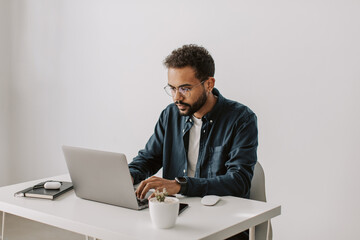 Portrait of young serious black businessman or student in glasses working at laptop in the office or at home sitting at the white table or desk. freelancer work from home, distant online job.