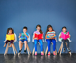 Lined up to learn. Studio shot of a group of kids sitting on chairs and reading books against a...