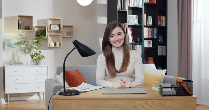Portrait shot of Caucasian young woman with long hair looking at camera and smiling joyfully at home. Beautiful happy female student sitting at table with laptop computer. Freelancer in cozy room