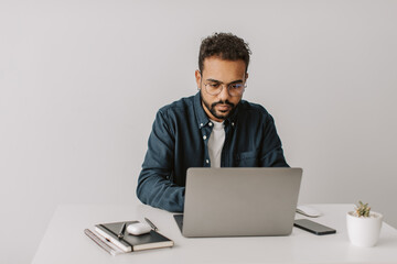 Attractive young african american diverse bearded man with curly hair in stylish shirt with glasses working on laptop sitting at home, remote job, work from home.