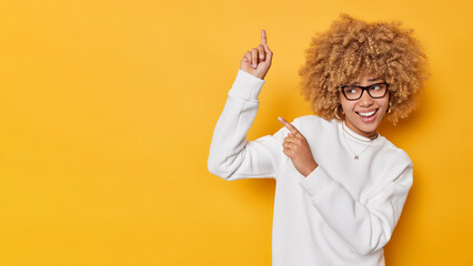 Cheerful curly haired adorable woman points above against blank space shows information banner demonstrates advertisement wears spectacles white casual jumper isolated over yellow background