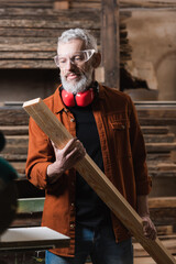 mature furniture designer in goggles holding wooden plank in carpentry studio.