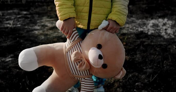 Kid holding fluffy bear with a burnt background after the bombing in Ukraine. during the war. Child in the colors of the Ukrainian flag pose with a toy bear. Stop War. Crisis, stop aggression, No War.