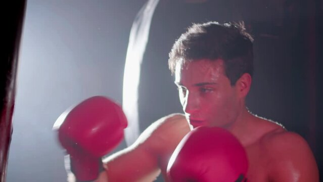 Strong male boxer hitting punching bag while working out in boxing gym alone. Active young man training indoors, practicing punches. Handheld shot. Low key. Sport, boxing concept