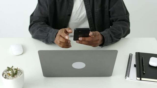 High angle view of one African american man typing and texting on smartphone sitting in front of laptop in white airy office, indoor black adult student study online, crypto enthusiast searching stock