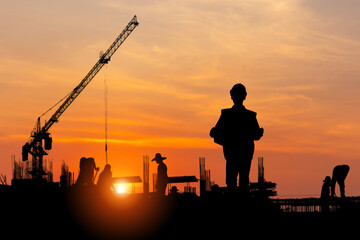 Silhouette of Engineer with blueprint and foreman worker team at construction site with blurred sunset sky background