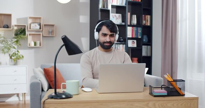 Arabian young handsome man in headphones sitting at desk, listening to music and working on laptop. Arab male typing on keyboard of computer in nice cozy room. Freelancer in home office.
