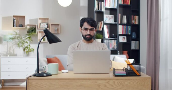 Arabian young handsome man in glasses sitting at desk and working on laptop. Arab male typing on keyboard of computer in nice cozy room. Freelancer in home office. Surfing online. Workday. E-studying.