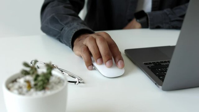 Close-up african-american mans hand working on wireless mouse in front of a laptop in a white modern home or office on a clean desk with glasses, green and office tech gadgets, slow motion 4k