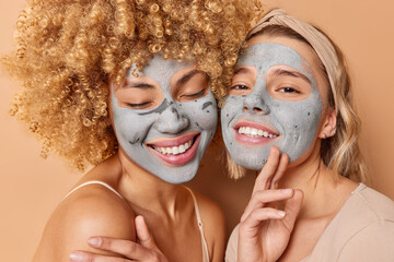Two happy friendly women stand closely to each other apply nourishing clay mask smile broadly wear headband dressed in casual clothes isolated over brown background. Beauty treatments concept