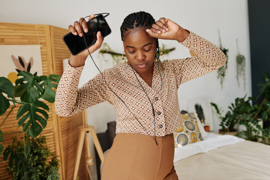African pregnant woman in headphones dancing in bedroom while listening to music on her mobile phone