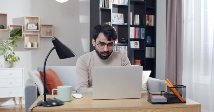 Young Arab handsome man in glasses working at laptop and getting good news or great result in work. At home. Arabian cheerful male freelancer sitting at computer. Gamer winning in game online.