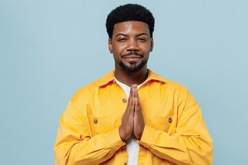 Obraz premium Young man of African American ethnicity 20s in yellow shirt hold hands folded in prayer gesture, begging about something isolated on plain pastel light blue background studio People lifestyle concept