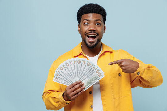 Young shocked amazed fun man of African American ethnicity 20s wear yellow shirt hold point index finger on fan of cash money in dollar banknotes isolated on plain pastel light blue background studio