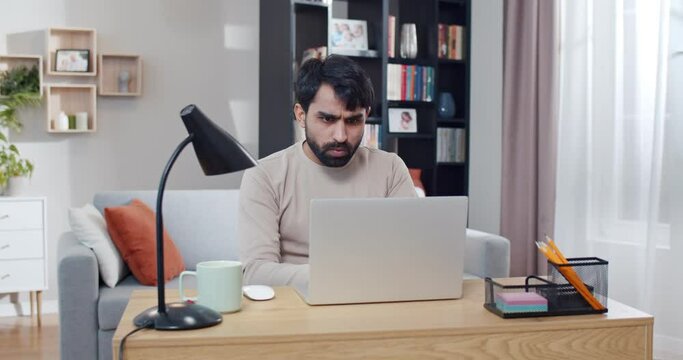 Young Arab handsome man working at laptop and getting good news or great result in job. At home. Arabian joyful male freelancer at computer. Gamer winning in game online. Lockdown concept.