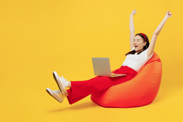 Full size young girl woman of Asian ethnicity 20s years old wear casual clothes sit in bag chair hold use work on laptop pc computer do winner gesture isolated plain yellow background studio portrait