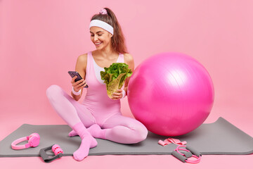 Full length shot of happy female fitness model checks calories via smartphone application holds green vegetable keeps to healthy diet poses on karemat with sport equipment against pink background