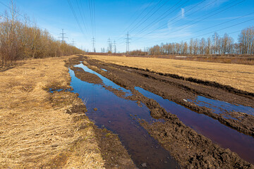 Rural dirt or unpaved road with mud and slush among  towers of electric main in the countryside field on the background of blue sky with clouds and the forest with the wires in sunny or spring weather
