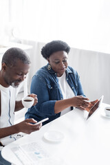 african american woman holding digital tablet near cheerful husband with cup of coffee using smartphone.