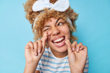 Positive woman with curly blonde hair wears headband and casual striped t shirt flossses teeth uses dental floss winks eye isolated over blue background. Oral hygiene and health care concept