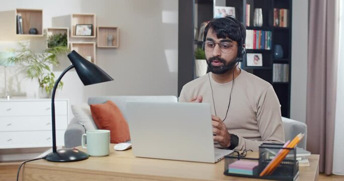 Arabian young man in headset working at computer and videochatting at home. Call center worker at home. Arab male having videochat at laptop. Freelancer work via webcam. Conversation.