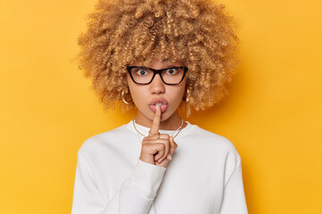 Surprised woman with curly bushy hair makes silence gesture keeps index finger over lips looks mysteriously wears transparent glasses and casual white jumper isolated over yellow background. - obrazy, fototapety, plakaty