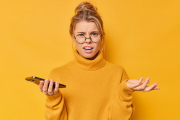 Upset disturbed dissatisfied young woman with fair hair gathered in bun raises palm shrugs shoulders wears round spectacles casual jumper holds cellular cannot understand how to send message