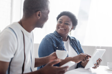 smiling african american woman pointing at digital tablet and looking at senior husband with cup of coffee.