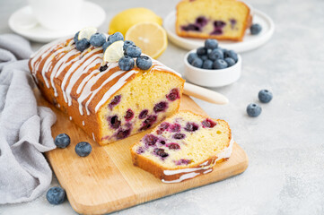 Lemon blueberry cake with lemon icing and fresh berries on top on the board on a gray concrete background with cup of tea. Selective focus. Copy space. - obrazy, fototapety, plakaty