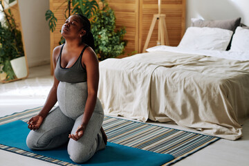 African pregnant girl sitting on exercise mat and meditating with her eyes closed in bedroom