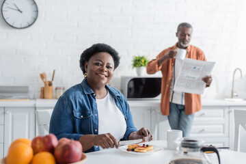 happy and senior african american woman having breakfast near blurred husband with newspaper.