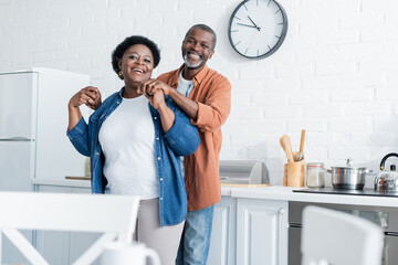 happy senior african american man holding hands of smiling wife in kitchen.