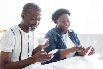 senior african american man with cup of coffee using smartphone near blurred wife with digital tablet.