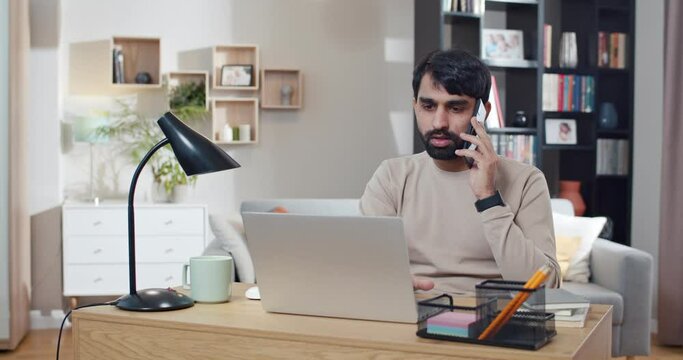 Young Arabian man speaking on mobile phone while working at laptop in cozy room. Arab businessman startupper talking on cellphone and sitting in front of computer at home. Freelancer. Conversation.