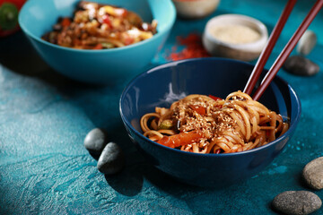 Japanese cuisine. Udon noodles with fried vegetables: carrots, eggplant, zucchini, soy sauce and sesame seeds in blue bowl on a turquoise background with fern leaves. Background image, copy space